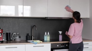 Woman in pink T-shirt wiping a white kitchen cabinet. Domestic cleaning in contemporary home environment