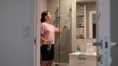 A woman sprays cleaning solution on a large glass window in a bathroom. Daily cleaning with an emphasis on hygiene. Glass cleaning with attention to detail and care for reflection