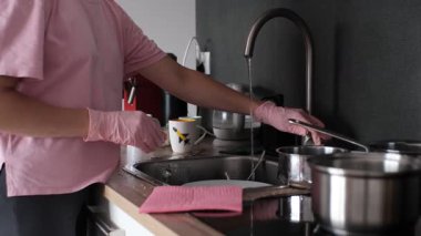 Woman in home outfit and gloves scrubs plate with sponge in bright modern kitchen 