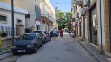 Kıbrıs Rum Kesimi 'nin tipik bir kentsel caddesi, parlak mavi gökyüzü altında yerli halkın yürüyüş ve günlük Akdeniz yaşamının yer aldığı park halindeki arabalar, dükkanlar ve balkonlarla dolu. Atmosfer şehir rutinini birleştiriyor