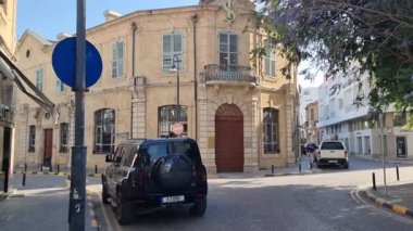 Güneşli bir sokak, eski Kıbrıs kasabasında kemerli kapıları ve ahşap panjurları olan görkemli tarihi bir taş binanın etrafında kıvrılıyor. Park edilmiş arabalar, yapraklı dallar ve ince şehir cazibesi...