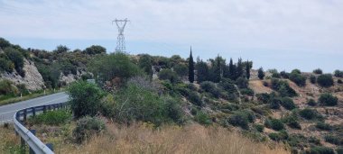 Akdeniz tarzı yamaçlarda kuru çim yamaçları ve yeşil çalılıklar boyunca kıvrılan metal korkulukları olan dolambaçlı bir kırsal yol. Uzakta, uzun selvi ağaçları karanlık bir küme oluşturur.
