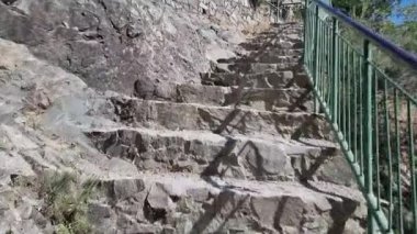 Kıbrıs dağlarında çalılar ve ağaçlarla çevrili kayalık bir uçurum duvarına tırmanan yeşil metal korkuluklu engebeli beton adımların videosu, zorlu yürüyüş yollarına güvenli bir gezinti olanağı sağlıyor.