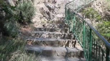 Kıbrıs dağlarında çalılar ve ağaçlarla çevrili kayalık bir uçurum duvarına tırmanan yeşil metal korkuluklu engebeli beton adımların videosu, zorlu yürüyüş yollarına güvenli bir gezinti olanağı sağlıyor.