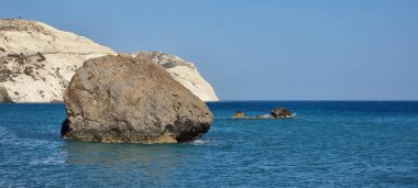 Derin mavi Akdeniz 'den yükselen toprak kırmızısı ve bej tonlarıyla yüksek, engebeli, beyaz kireçtaşı kayalıklardan oluşan panoramik bir manzara. Kayanın üzerinde belirgin büyük gri bir kaya parçası duruyor.