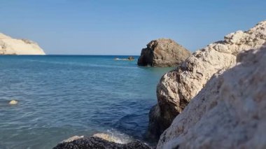 Kıbrıs 'ın Afrodit Sahili yakınlarındaki turkuvaz Akdeniz' de kayalık büyük bir çıkıntı, arka planda açık mavi gökyüzü altında beyaz kireçtaşı kayalıkları ve tepeler, mitoloji ve kıyı için mükemmel.