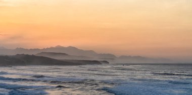 Gün batımında vahşi okyanus kıyıları. Dalgalar kırılıyor ve dağlar siluet içinde.