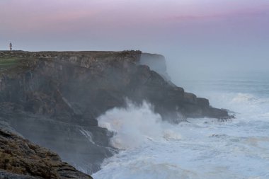 İspanya 'daki Cape Ajo' ya büyük fırtına dalgaları vuruyor. Güneş doğarken yukarıdaki kayalıklarda deniz feneri var.