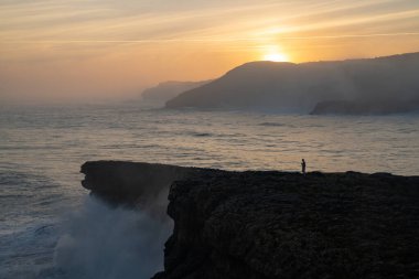 Büyük bir fırtınanın dalgalanan okyanus dalgalarının kıyıya vuruşunu ve gün doğumunda uçurumların orada dikilen bir insanın görüntüsü.