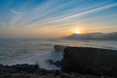 Büyük bir fırtınanın dalgalanan okyanus dalgalarının kıyıya vuruşunu ve gün doğumunda uçurumların orada dikilen bir insanın görüntüsü.