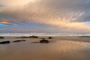 Güzel, boş bir kumsalda gün doğumu dalgalar çarpıyor ve kumlarda kayalar var.