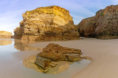 Sıcak akşam ışığında gelgit havuzları ve pürüzlü uçurumları olan kumlu bir sahil manzarası.