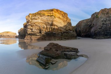Sıcak akşam ışığında gelgit havuzları ve pürüzlü uçurumları olan kumlu bir sahil manzarası.