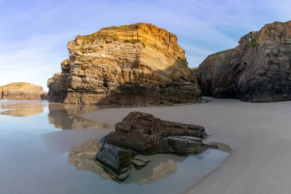 Sıcak akşam ışığında gelgit havuzları ve pürüzlü uçurumları olan kumlu bir sahil manzarası.