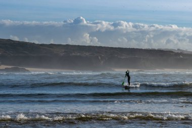 Vila Nova de Milfontes, Portekiz: 23 Aralık 2020: Sörfçü Milfontes plajında bir SUP küreğinde dalgaları yakalamayı seviyor