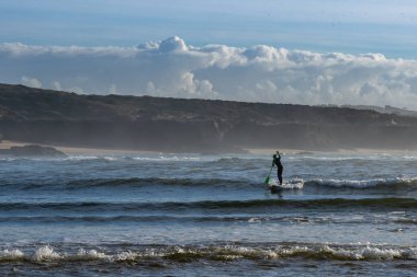 Vila Nova de Milfontes, Portugal: 23 December 2020: surfer enjoys catching waves on a SUP paddleboard at Milfontes beach clipart