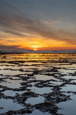 Okyanusun üzerinde kayalık plajlı ve ön planda gelgit havuzlu güzel bir gün batımının dikey görüntüsü.