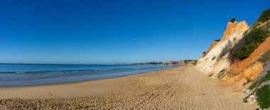 Güneşli bir günde renkli kum kayalıkları olan geniş ve boş bir kumsal manzarası.