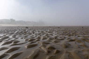 Sis tabakası, dalgaların çekildiği uçsuz bucaksız bir deniz sahilinin üzerinden yükseliyor.