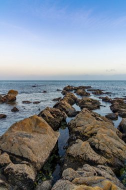 Gün batımında sakin bir okyanusa açılan kahverengi taşlı huzurlu bir deniz manzarası.