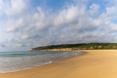 Huzurlu kumlu sahil, yumuşak dalgalar ve arka planda ağaçlarla kaplı tepeler ve yukarıda beyaz kabarık bulutlar.