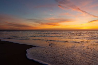 Okyanus ve sahil üzerinde muhteşem renkli bir gün batımı. Kıyıda hafif dalgalar çakıyor.