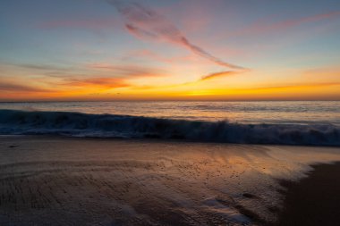 Okyanus ve sahil üzerinde muhteşem renkli bir gün batımı. Kıyıda hafif dalgalar çakıyor.