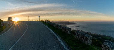Güneş doğarken deniz kıyısında bir dağın tepesine park etmiş bir karavan. Kıyıyı ve okyanusu harika görüyor.