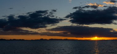 Okyanus üzerinde manzaralı ve renkli bir gün batımı dalgaların üzerinde parıldayan ışık ve arkadaki dağların silueti.