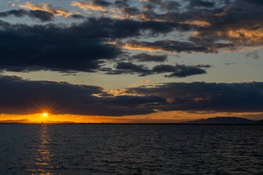 Okyanus üzerinde manzaralı ve renkli bir gün batımı dalgaların üzerinde parıldayan ışık ve arkadaki dağların silueti.