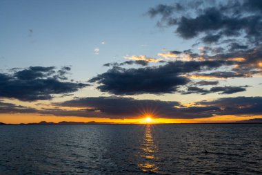 Okyanus üzerinde manzaralı ve renkli bir gün batımı dalgaların üzerinde parıldayan ışık ve arkadaki dağların silueti.