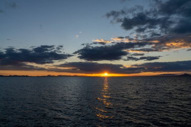 Okyanus üzerinde manzaralı ve renkli bir gün batımı dalgaların üzerinde parıldayan ışık ve arkadaki dağların silueti.