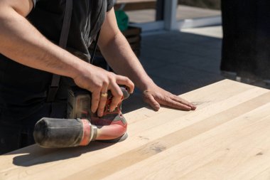 Büyük ahşap bir masayı zımparalamak için kablosuz zımpara makinesi kullanan bir inşaat işçisi.