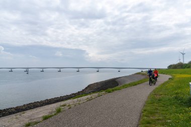 Collijnsplaat, Hollanda - 13 Mayıs 2021: Aile, arka planda Zeelandbrug ile birlikte Doğu Şehidi Mahallesi boyunca bisiklet sürmekten keyif alıyor.