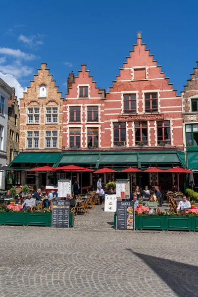 Bruges, Belçika - 12 Mayıs 2021: İnsanlar Brüksel 'deki Pazar Meydanı' ndaki restoranlarda, arka planda birçok tarihi tuğla bina bulunan bir günün tadını çıkarıyorlar.