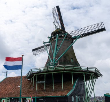 A traditional Duthc windmill under an overcast sky with the flag of the Netherlands flying in a strong wind