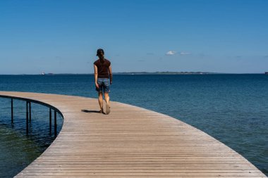 Sıska bir kadın, durgun mavi bir okyanusun üzerinde dairesel bir sahil yolunda yürüyor.
