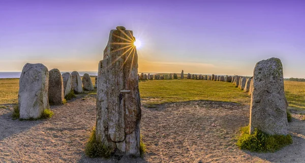 Landscape view of the prehistoric Ales Stenar ship setting on the southern coast of Sweden at sunset under a lilac sky with a sun star