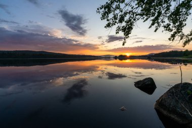 Sakin bir gölün üzerinde renkli bir günbatımı. Suda yansımaları var.