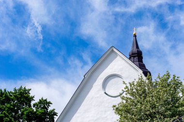 Aşağıda beyaz bulutlar ve yeşil ağaçlar olan mavi gökyüzü altında çan kulesiyle bembeyaz bir kilise binasının detaylı bir görüntüsü.