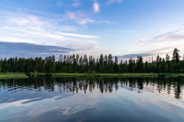 Çam ağaçlarıyla kaplı bir kıyı manzarası ve suya yansıyan anlamlı bir gökyüzü.