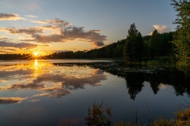 Renkli bir günbatımı, yeşil orman ve sazlıklar ile sakin bir göl manzarasına yansıyor.