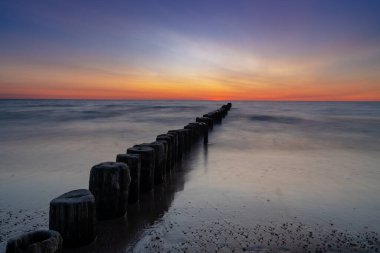 Okyanus günbatımının kumlu sahili ve ön planda ahşap fırtına kasığı olan uzun pozlama manzarası.