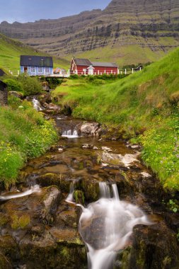 Faroe Adaları 'ndaki Kunoy köyünde huzurlu bir şelalenin ve renkli ahşap evlerin dikey görüntüsü