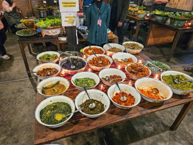 Kapalı pazar ortamındaki ahşap bir masada çeşitli çanaklar ve servis aletleriyle sergilenen çeşitli tabaklar..