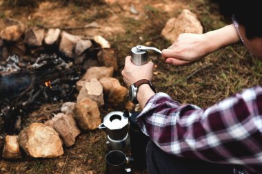 Kahve molası vermek, kamp ateşinde kahve öğütücü elle işlemek, Turizm Kampı Gezisi, Yürüyüş Macerası ve Turizm Ekipmanları Konsepti.