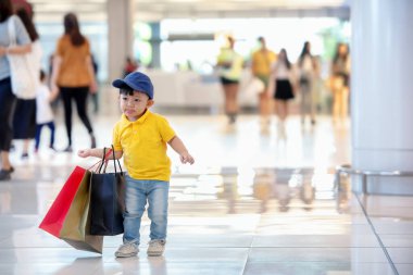 Çocuk alışverişi. Sarı tişörtlü ve kot pantolonlu Asyalı küçük çocuk dükkanın keyfini çıkarıyor. Alışveriş merkezinde ağır alışveriş torbalarıyla dolaşmak..