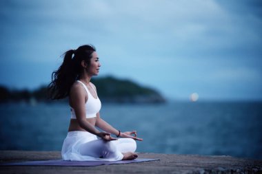 Sukhasana egzersizinde yoga yapan Asyalı genç bir kadın denize bakarken....