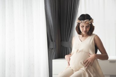 Hamilelik, hamile, karnında hamile bir kadın. Evde pencerenin yanında oturan hamile elbiseleri. Annelik hazırlığının mutluluğu ve fotokopi alanı konseptiyle beklenti.