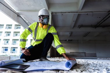 İnşaat işçisi yüz maskesi ve güvenlik kaskı takıp inşaat alanındaki çizimleri takip eden bir bölgeye bakıyor. İnşaat Mühendisliği ya da İnşaat Mühendisliği yıkılan binayı kontrol ediyor..